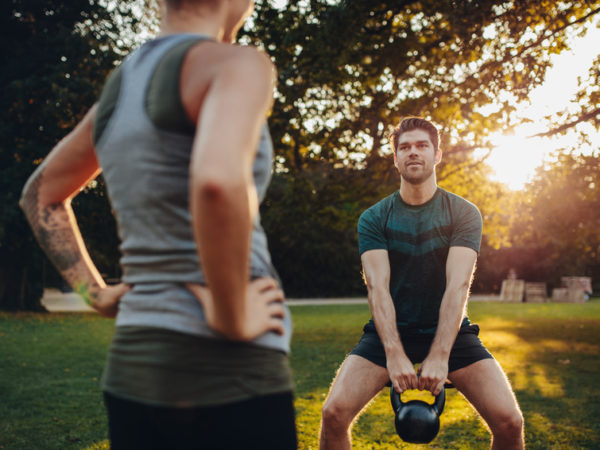 Fitness man exercising with personal trainer Young man doing weight training with personal female trainer in the park. Caucasian male exercising with kettlebell.