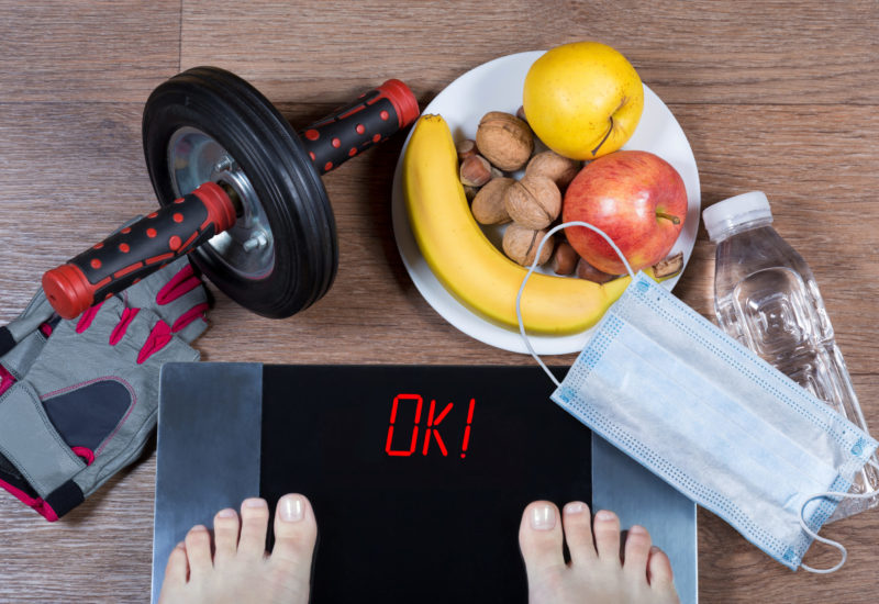 Girl checks her weight after quarantine. Digital scales with word ok surrounded by sport accessories, healthy food, water bottle and face mask. Concept of healthy lifestyle during self-isolation.